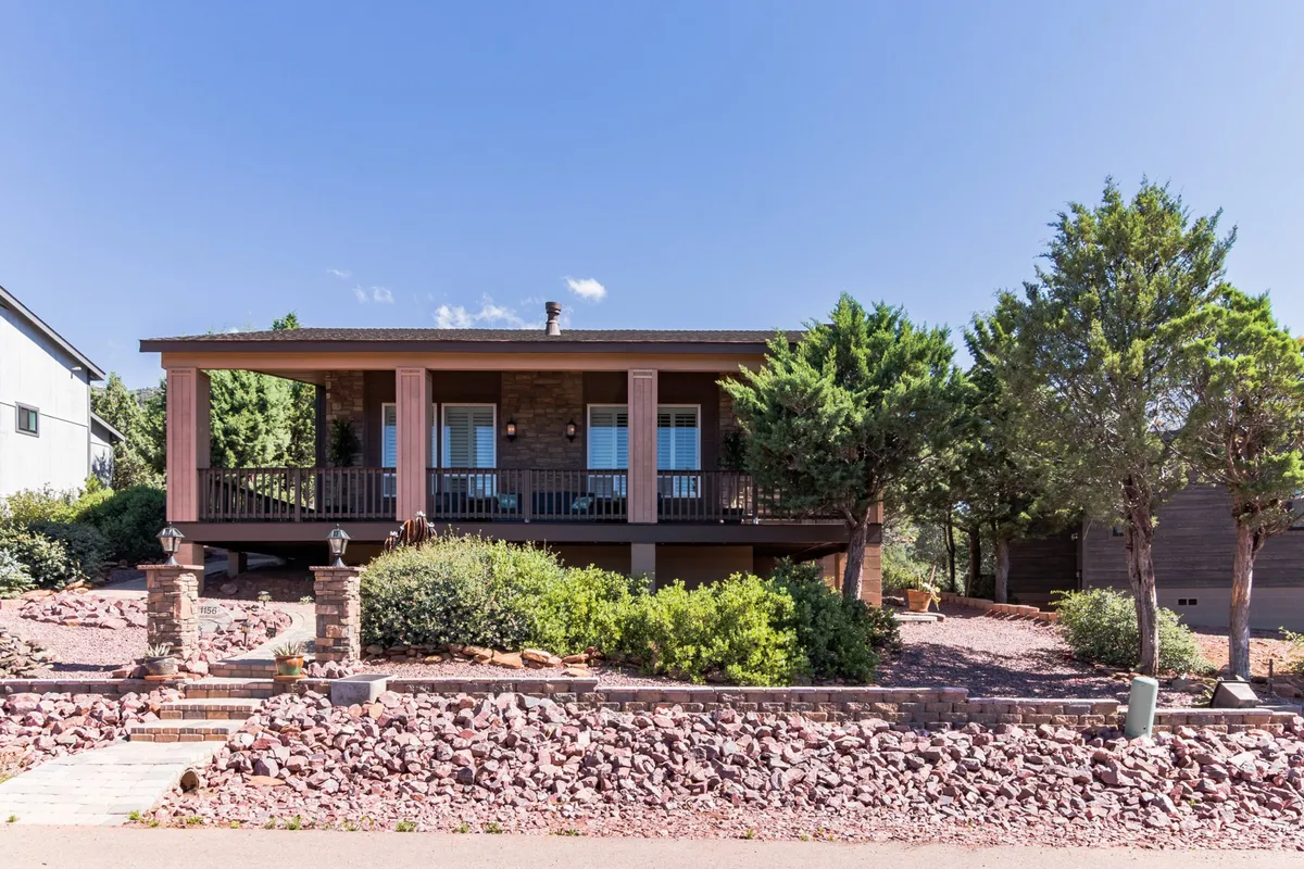 A welcoming front porch surrounded by a landscaped rock garden and green shrubs, with sturdy stone pillars supporting the structure. The porch looks ideal for relaxing, socializing, or enjoying the view, with ample space for both people and pets to unwind together.