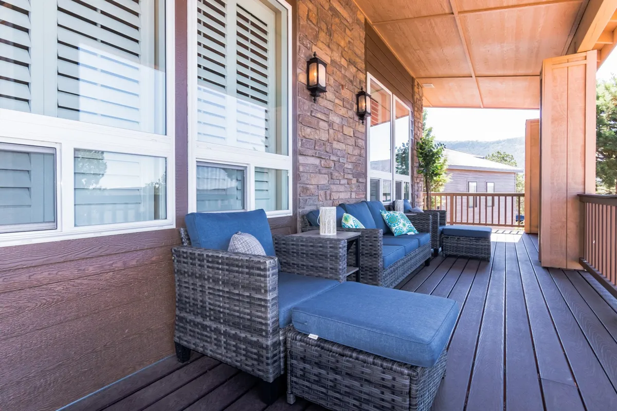 An exterior shot of a wooden deck with comfortable outdoor seating, decorative cushions, and a wooden railing set against a lush green backdrop. The setting is casual and welcoming, perfect for morning coffee, socializing, or simply enjoying the sights and sounds of nature.