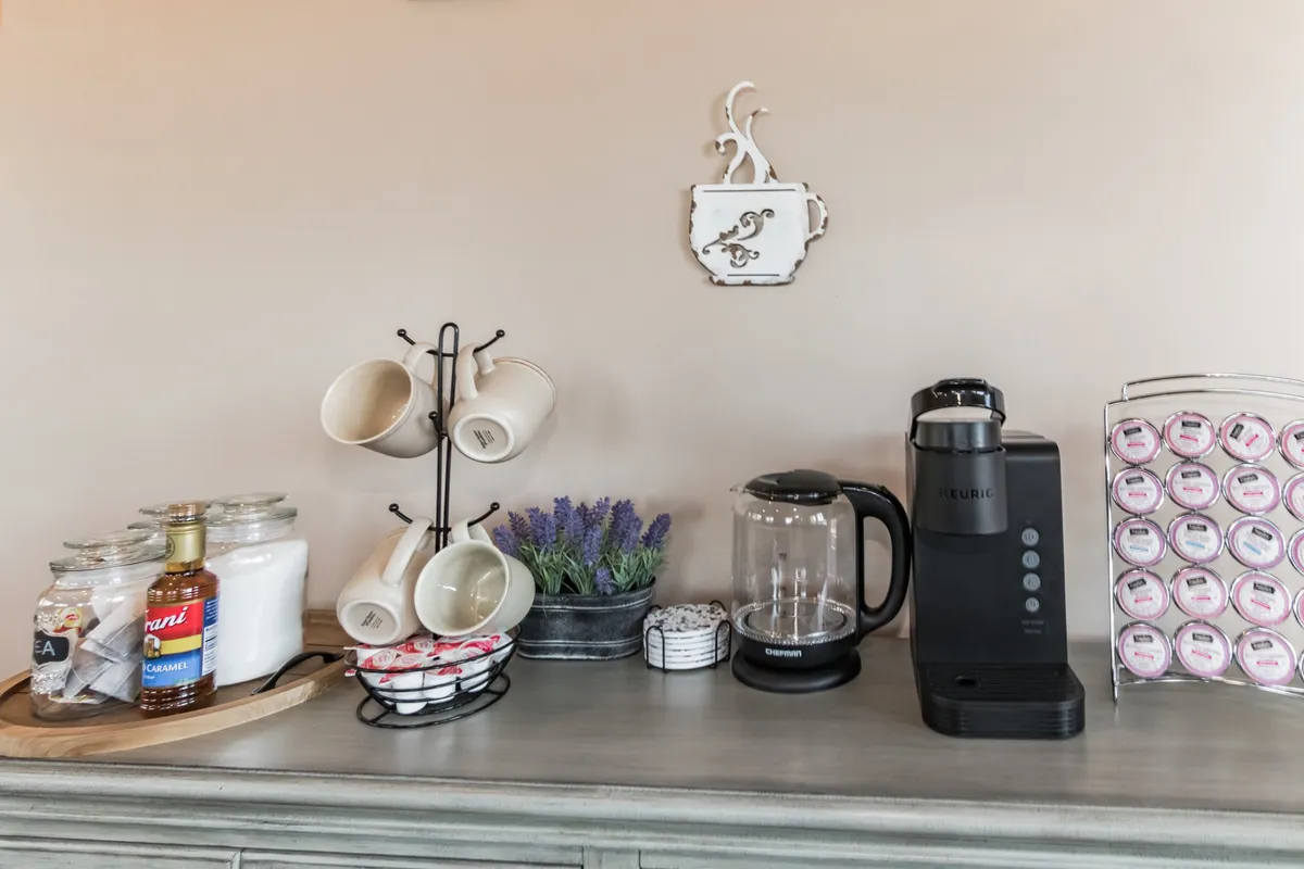 A cozy beverage nook featuring a coffee machine, an assortment of mugs, tea jars, and lavender plants. The space exudes warmth and is clearly designed for morning routines or relaxing with a hot drink.