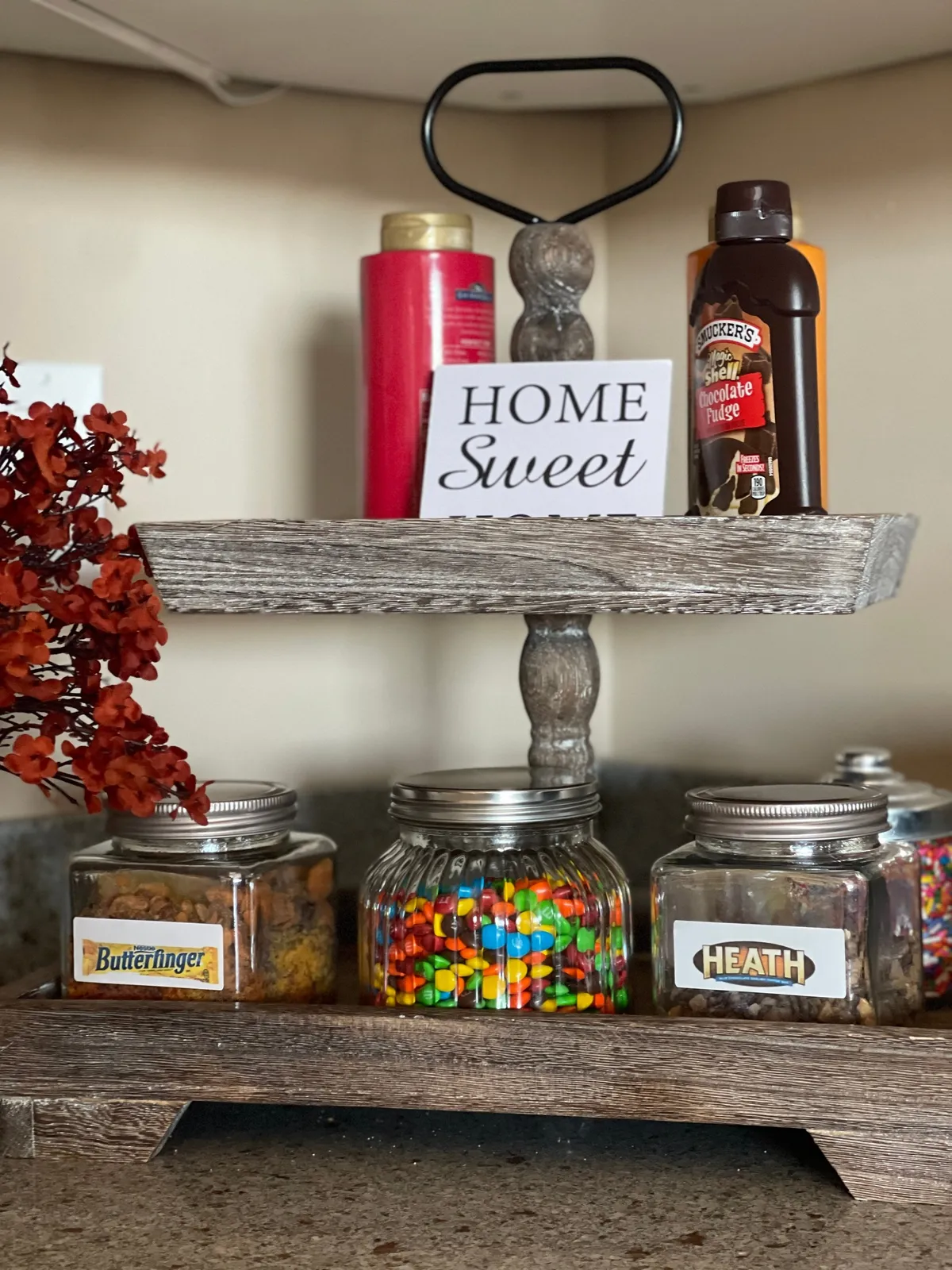 A wooden display stand adorned with jars of colorful candies, a 'HOME Sweet' sign, and decorative flowers. The setup is playful and inviting, evoking the joy of sharing sweet treats.
