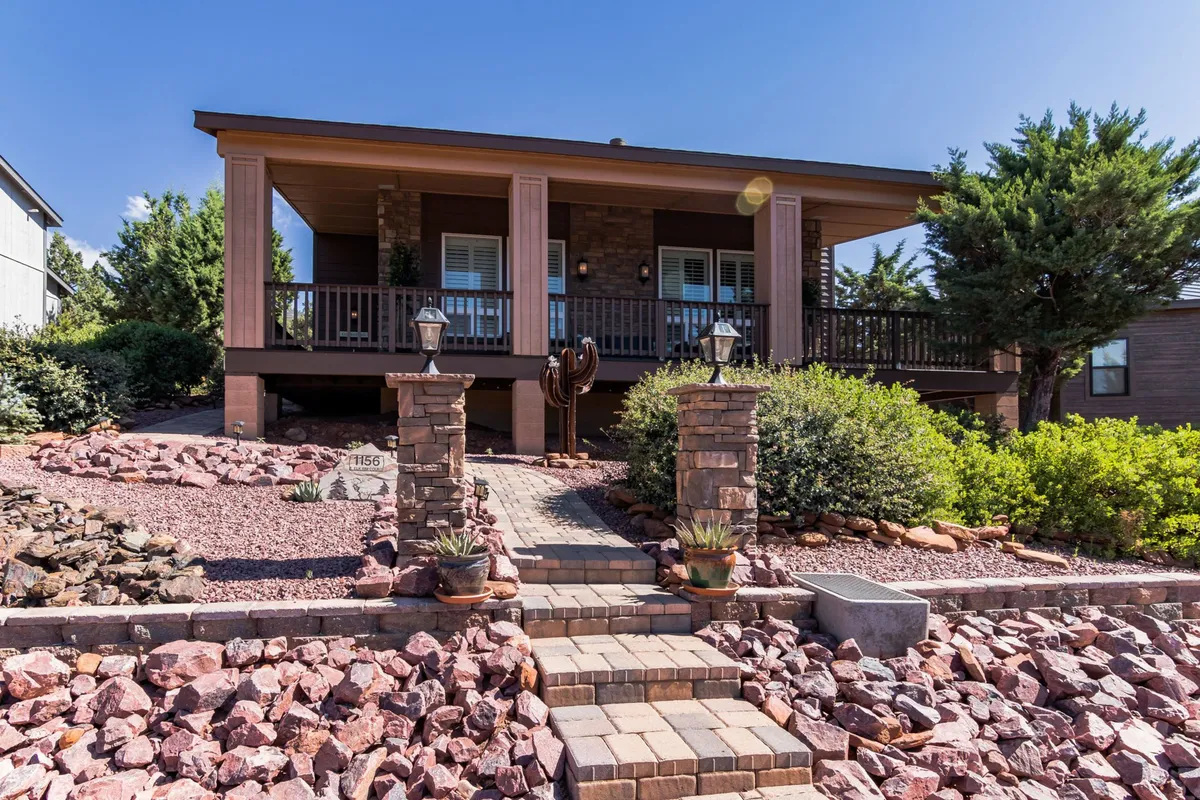 A serene exterior shot featuring a landscaped garden, stone pathway, inviting front porch, rock formations, and decorative lamp posts surrounded by greenery. The porch is the ideal spot to unwind after a day outdoors.